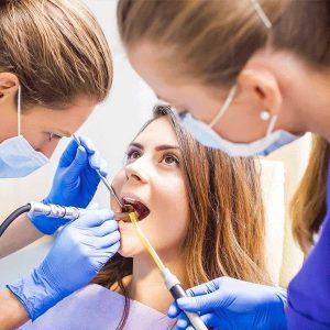 Dental professional in blue gloves and mask examines patient's teeth in a bright clinical setting